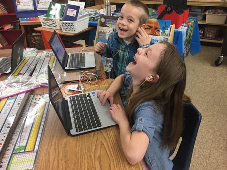 A boy and a girl in a classroom using laptops laughing